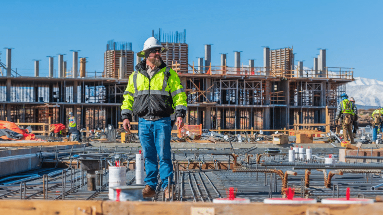 A person in a safety jacket and hardhat standing in front of a construction site.